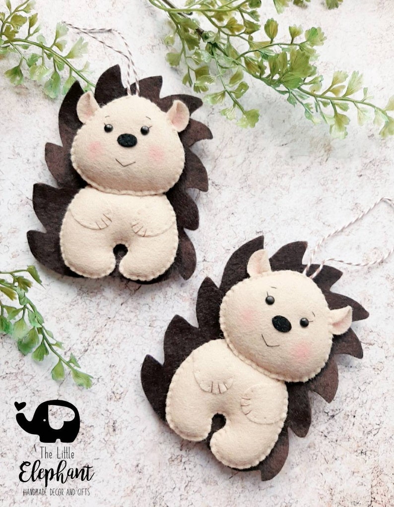May include: Two felt hedgehog ornaments with brown spines and cream-colored bodies. Each has a smiling face with pink cheeks, black eyes, and a black nose. They hang from white string. The Little Elephant logo is in the lower left.