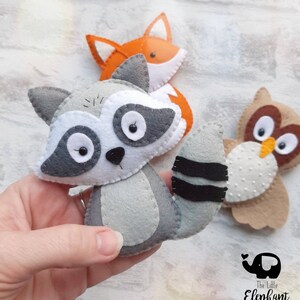 May include: A set of three felt animal ornaments, including a raccoon, a fox, and an owl. The raccoon is gray with black eyes and a black and white striped tail. The fox is orange with a white chest and a black nose. The owl is brown with white spots and yellow eyes. The ornaments are handmade and would be a great addition to any nursery or child's room.
