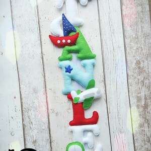 May include: A felt letter banner spelling the name "STANLEY" with a variety of colorful vehicles and nautical designs. The letters are white, blue, green, and red. The vehicles include a red car, a green car, a white airplane, and a red sailboat. The banner is hanging on a white wooden background.