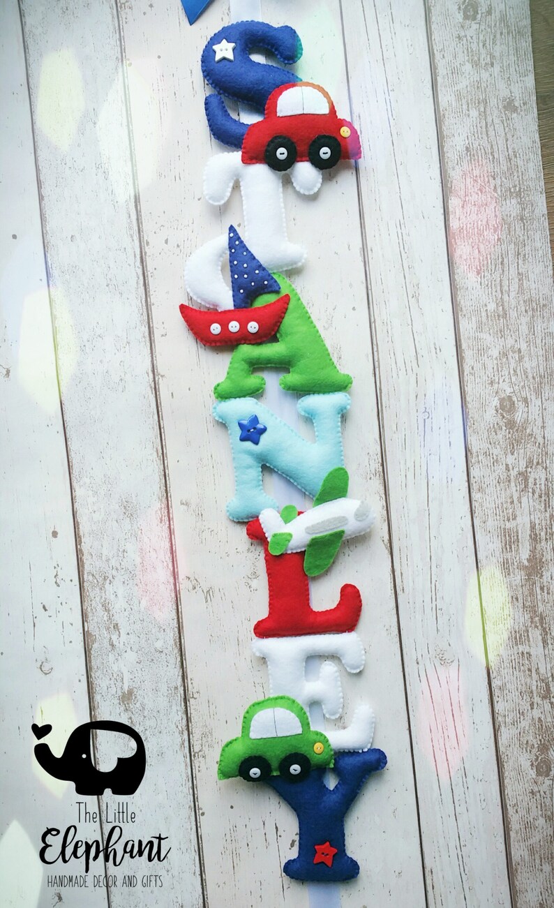 May include: A felt letter banner spelling the name "STANLEY" with a variety of colorful vehicles and nautical designs. The letters are white, blue, green, and red. The vehicles include a red car, a green car, a white airplane, and a red sailboat. The banner is hanging on a white wooden background.