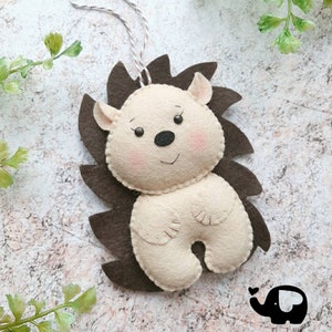May include: A handmade felt hedgehog ornament with a brown spiky back and a cream-colored body. The hedgehog has a smiling face with pink cheeks, small ears, and a black nose. A string is attached for hanging.