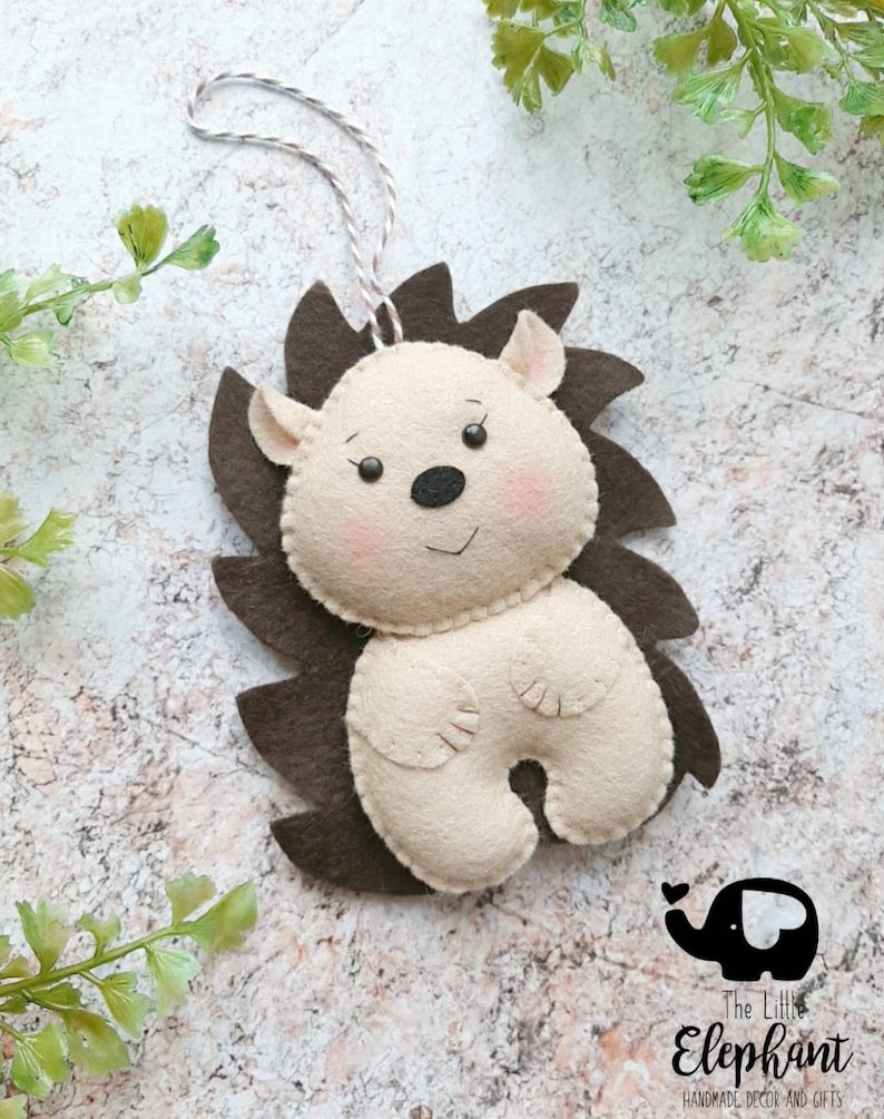 May include: A handmade felt hedgehog ornament with a brown spiky back and a cream-colored body. The hedgehog has a smiling face with pink cheeks, small ears, and a black nose. A string is attached for hanging.