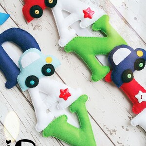 May include: Felt letter banner with the name "Davy" in white, green, and blue letters. Each letter has a small felt car attached to it in red, blue, and light blue. The cars have black wheels and yellow headlights. The banner is hanging on a white wooden background.