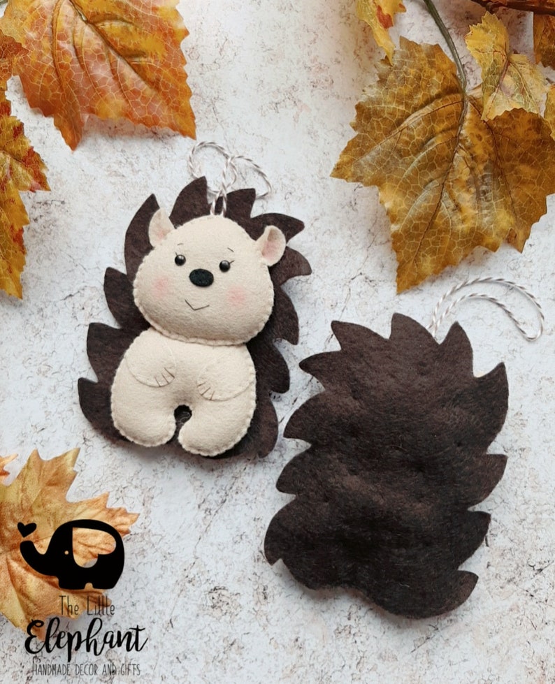 May include: Two felt hedgehog ornaments. One is complete with a cream-colored body, brown spikes, and a smiling face. The other is a brown felt spike shape. Both have a silver string for hanging. The Little Elephant logo is in the bottom left.
