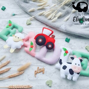 May include: A felt name banner with the name "EAZIE" spelled out in green, pink, and white felt. The letters are decorated with felt flowers, a sheep, a tractor, and a cow. The banner is hanging from a string and the word "play" is written on a wooden sign below.