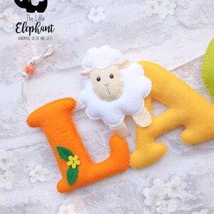 May include: A felt banner with the letters "LA" in orange and yellow. The letter "L" has a green leaf with a yellow flower. A white felt sheep is attached to the letter "A". The banner is handmade and has a white background.