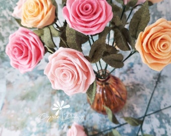May include: A bouquet of felt roses in shades of pink and peach, arranged in a clear glass vase with a brown base. The roses are crafted with multiple layers of felt, creating a realistic and delicate appearance. The bouquet is a charming and unique decorative piece.