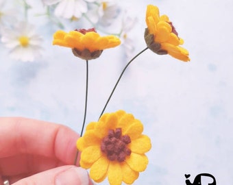 May include: Three felt sunflowers with brown centres on wire stems. The sunflowers are yellow and the centres are a dark brown. The flowers are held in a hand.