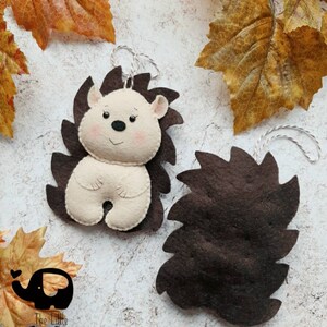 May include: Two felt hedgehog ornaments with brown spines and cream-colored bodies. The hedgehogs have small black eyes, pink cheeks, and a string for hanging. The Little Elephant logo is in the lower left corner.