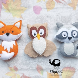 May include: Three felt animal ornaments: a fox with an orange body and white tail, an owl with brown and white feathers, and a raccoon with gray fur and a black and white striped tail.