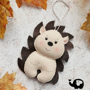 May include: A handmade felt hedgehog ornament with a cream-colored body, brown spines, and a string for hanging. The hedgehog has a smiling face with pink cheeks and small ears. The Little Elephant logo is in the bottom right corner.