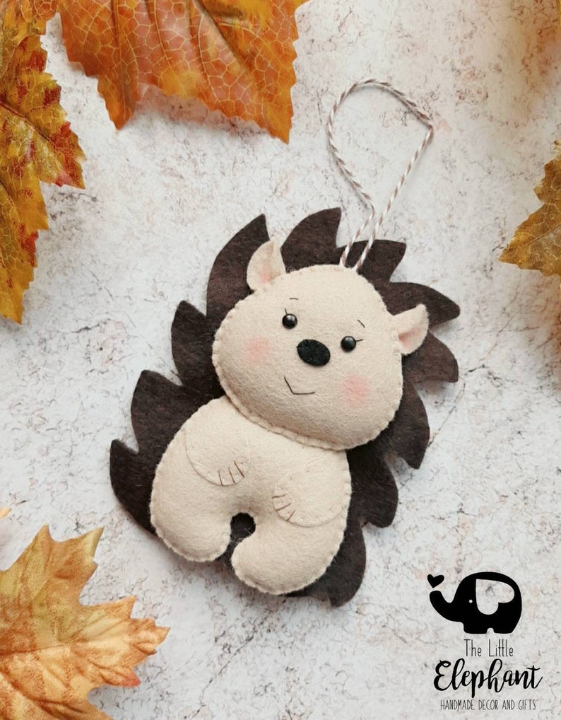 May include: A handmade felt hedgehog ornament with a cream-colored body, brown spines, and a string for hanging. The hedgehog has a smiling face with pink cheeks and small ears. The Little Elephant logo is in the bottom right corner.