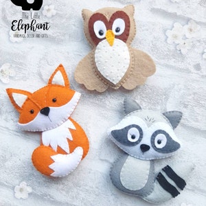 May include: Three felt animal ornaments: a brown owl, an orange fox, and a gray raccoon. The owl has large white eyes and a yellow beak. The fox has a white chest and tail tip. The raccoon has a black mask and a striped tail.