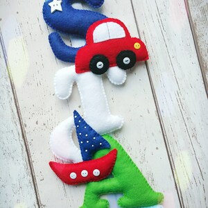 May include: A felt letter banner with the name "SIAN" spelled out in blue, white, green, and light blue felt. Each letter has a different design element, including a car, a sailboat, and a star. The banner is hung on a white wooden background.