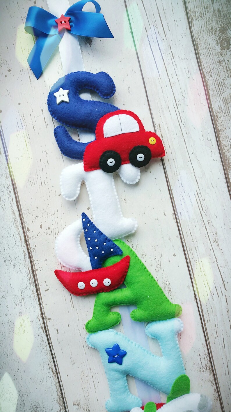May include: A felt letter banner with the name "SIAN" spelled out in blue, white, green, and light blue felt. Each letter has a different design element, including a car, a sailboat, and a star. The banner is hung on a white wooden background.