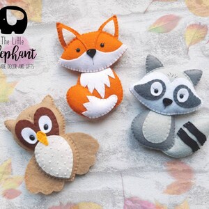 May include: Three felt animal ornaments: a brown owl with white spots, an orange fox with a white belly, and a gray raccoon with black stripes. The animals are handmade and have embroidered details. The text "The Little Elephant Handmade Decor and Gifts" is visible in the top left corner.