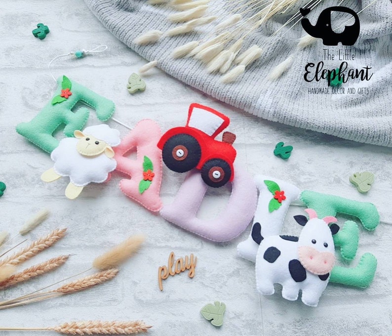 May include: A felt name banner with the name "EAZIE" spelled out in green, pink, and white felt. The letters are decorated with felt flowers, a sheep, a tractor, and a cow. The banner is hanging from a string and the word "play" is written on a wooden sign below.