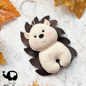 May include: A handmade felt hedgehog ornament with a brown spiky back and a cream-colored body. The hedgehog has a smiling face with pink cheeks and a black nose. A string is attached for hanging. The Little Elephant logo is in the bottom left corner.