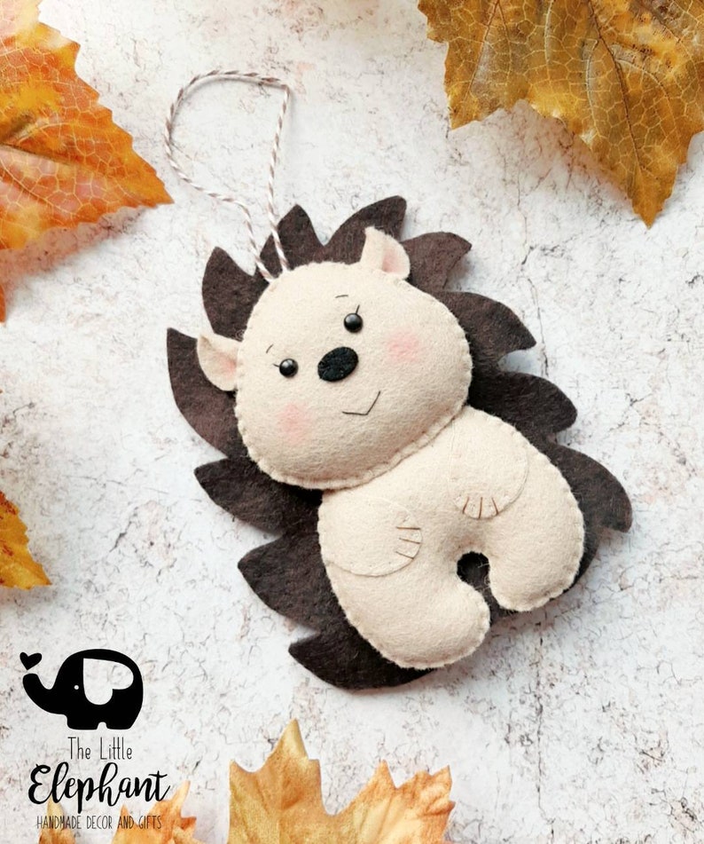May include: A handmade felt hedgehog ornament with a brown spiky back and a cream-colored body. The hedgehog has a smiling face with pink cheeks and a black nose. A string is attached for hanging. The Little Elephant logo is in the bottom left corner.