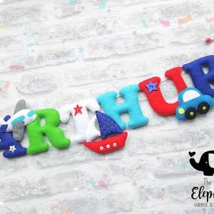 May include: A felt letter banner with the name "ARTHUR" in colorful letters. Each letter has a small felt appliqu&eacute; of a plane, boat, car, or star. The banner is strung on a white cord with red beads. The banner is on a white brick background. The text "The Little Elephant Handmade Decor and Gifts" is in the bottom right corner.