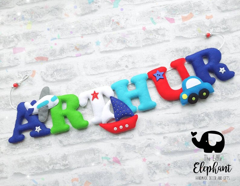 May include: A felt letter banner with the name "ARTHUR" in colorful letters. Each letter has a small felt appliqu&eacute; of a plane, boat, car, or star. The banner is strung on a white cord with red beads. The banner is on a white brick background. The text "The Little Elephant Handmade Decor and Gifts" is in the bottom right corner.