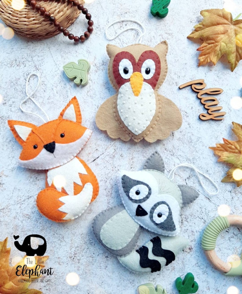 May include: Three felt animal ornaments: a brown owl with orange beak and white eyes, an orange fox with white accents, and a gray raccoon with black eyes and stripes. The ornaments are hanging from white strings and are surrounded by fall leaves and other decorative elements. The text "play" is written on a wooden sign.