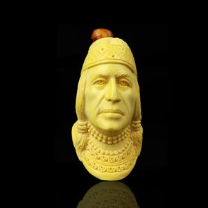 May include: A carved, yellow-toned bust of a person with long hair, a headdress, and a beaded necklace. The bust is set against a black background, with a small, brown object above the head. The carving has detailed facial features and decorative elements.