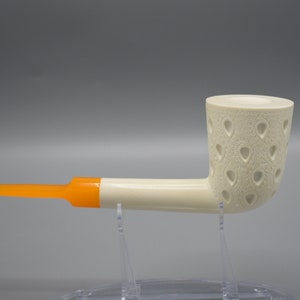 May include: A white smoking pipe with an orange stem and a patterned bowl. The bowl has a design of small, teardrop-shaped holes.