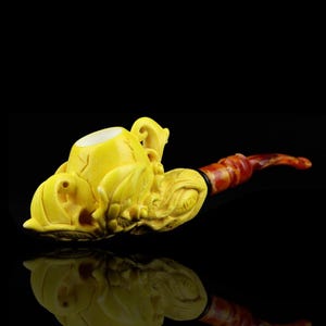 May include: A yellow carved tobacco pipe with an intricate design. The bowl is a light yellow, and the stem is reddish-brown with a dark band near the bowl. The pipe is set against a black background.