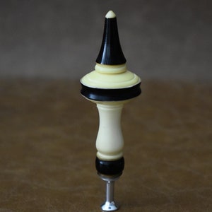 May include: A white and black decorative bottle stopper with a pointed top and a silver screw base.
