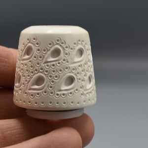 May include: Ivory-colored thimble with a decorative pattern of teardrop shapes and small circles. The thimble has a rounded top and a flared base. The design is carved into the surface, creating a textured effect. The thimble is held in a hand.