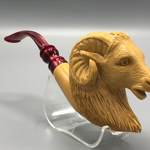 May include: A wooden pipe carved in the shape of a ram's head with a red acrylic stem. The pipe is displayed on a clear acrylic stand.