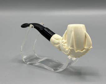 May include: Ivory-colored tobacco pipe with a black stem and mouthpiece. The bowl is carved with a floral design. The pipe is displayed on a clear acrylic stand against a gray background.