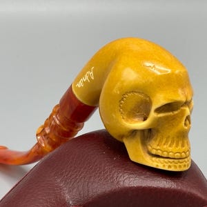 May include: A yellow skull-shaped pipe with a red and yellow stem. The pipe is engraved with the word "Altan".