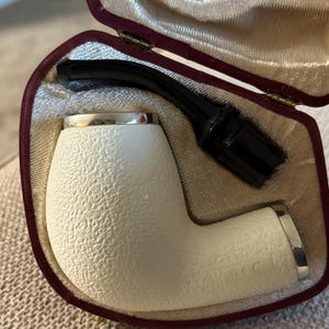 May include: A white pipe with a textured surface and silver accents, presented in a burgundy velvet case. The pipe has a black stem and is likely for smoking tobacco. The case is open, revealing the pipe's components.