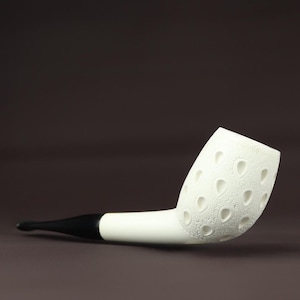 Pode incluir: Um cachimbo branco com um bocal preto. A tigela apresenta um padrão decorativo com recortes em forma de gota. O cachimbo está sobre um fundo castanho.