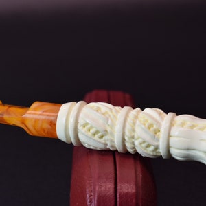 May include: A white carved cigarette holder with an orange amber mouthpiece. The holder has a detailed design of interwoven rope and floral patterns.