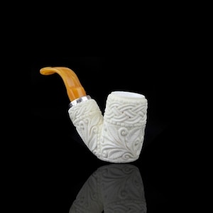 May include: Ivory-colored tobacco pipe with intricate floral and woven pattern carvings. The stem is a light amber color, and the pipe has a silver band. The pipe is set against a black background.