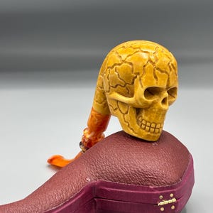 May include: A carved tobacco pipe with a skull-shaped bowl and a stem. The pipe is a light tan color with a textured surface. It rests on a burgundy leather case with gold-colored clasps. The stem is orange and yellow.