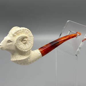 May include: A white ceramic pipe shaped like a ram's head with a red and orange stem.