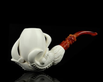 May include: A white tobacco pipe with a detailed, carved design. The bowl is a smooth, rounded shape with two handles. The stem is a reddish-brown color with a curved design. The pipe is resting on a reflective black surface.