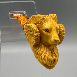 May include: A hand-carved wooden pipe with a ram's head design. The pipe is made of light brown wood and has a detailed carving of a ram's head with horns. The pipe is a unique and decorative piece.
