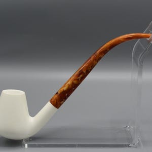 May include: A tobacco pipe featuring a smooth, matte white bowl and a long, curved stem with a marbled brown and amber pattern. The pipe is supported by a clear acrylic display stand, showcasing the contrast between the solid white and the swirl.