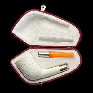 Pode incluir: Uma caixa de couro sintético vermelha aberta revela um interior branco, contendo um cachimbo branco com um padrão decorativo e um bocal laranja e prateado. A caixa é fechada com um fecho dourado.