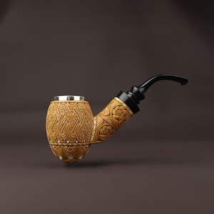 May include: A carved tobacco pipe with a light brown bowl and stem, featuring intricate floral and geometric patterns. The mouthpiece is black, and the pipe has a silver rim. The pipe is set against a dark brown background.