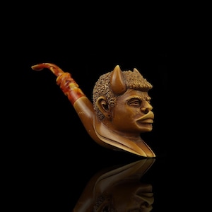 May include: A wooden pipe with a carved human head with horns. The pipe is brown and the head is painted in shades of brown and black.