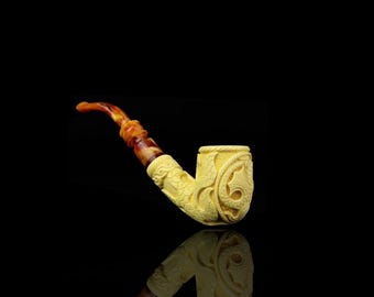 May include: A cream-colored, carved tobacco pipe with a curved stem and bowl. The stem is a mix of amber and brown hues. The bowl and stem are intricately carved with floral and geometric patterns. The pipe is set against a black background.
