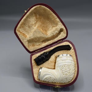 May include: An ivory-colored tobacco pipe with intricate floral carvings, a black stem, and silver accents, presented in an open burgundy velvet-lined case. The case has a gold clasp.