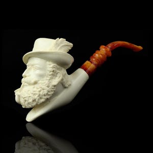 May include: A cream-colored tobacco pipe featuring a sculpted face of a man with a beard and hat. The pipe's stem is a light brown color, and the bowl is shaped like a man's head. The pipe is set against a black background.