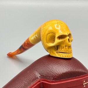 May include: A yellow skull-shaped pipe with a red and orange swirl design on the stem. The pipe is sitting on a red leather case with the word "Adan" inscribed on the stem.